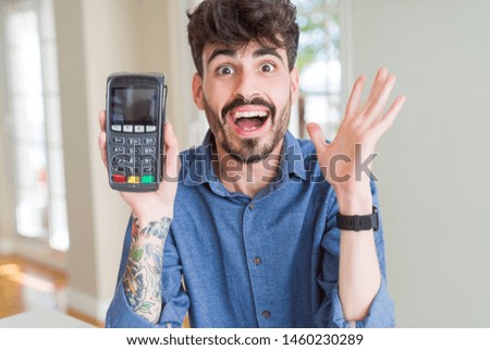 Young man holding dataphone point of sale as payment very happy and excited, winner expression celebrating victory screaming with big smile and raised hands