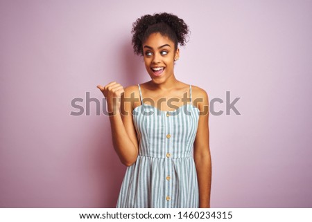 African american woman wearing casual striped dress standing over isolated pink background smiling with happy face looking and pointing to the side with thumb up.