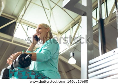 Low angle view of disabled active senior Caucasian woman with leg amputee talking mobile phone in fitness studio. Strong active senior female amputee training and working out