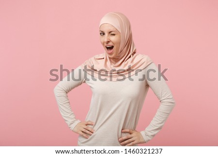Stunning young arabian muslim girl in hijab light clothes posing isolated on pink background. People religious Islam lifestyle concept. Mock up copy space. Blinking standing with arms akimbo on waist