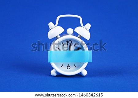 White alarm clock on blue background. Blank sticky note on the alarm clock for text. Reminder. Space for copy. Minimal concept.
