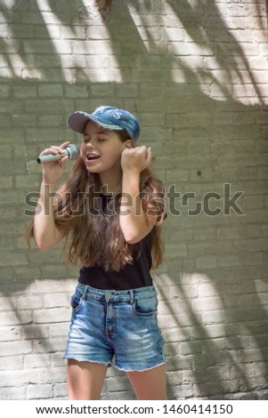 Beautiful little girl with microphone on brick wall background.The girl in denim shorts sings into a microphone on a city street. Beautiful brunette. Vote. Long hair in a child. Lifestyle