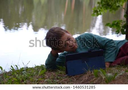 Boy schoolboy with a tablet in a beautiful meadow near the lake plays games, watches cartoons. Boy Child playing with Tablet PC Outdoor with forest on background Computer Game Dependence concept