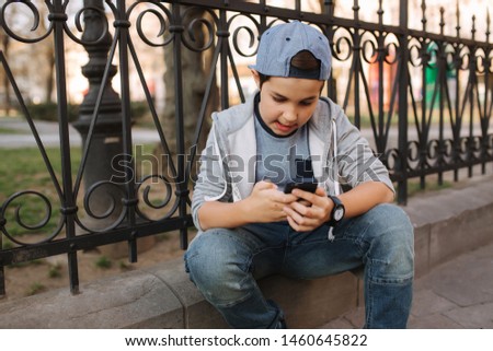 Young boy play online game at smartphone outside. Boy sitting in centre of the city. Boy play games while his mom going to the shop