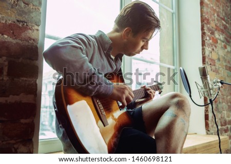 Young man recording music video blog, home lesson or song, playing guitar or making broadcast internet tutorial while sitting in loft workplace or at home. Concept of hobby, music, art and creation.