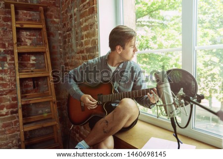 Young man recording music video blog, home lesson or song, playing guitar or making broadcast internet tutorial while sitting in loft workplace or at home. Concept of hobby, music, art and creation.