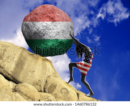 The girl, wrapped in the flag of the United States of America, raises a stone to the top in the form of a silhouette of Hungary