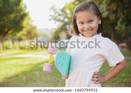 Happy cute little Asian girl smiling to the camera confidently, holding her mint blue penny board, copy space. Cheerful little girl enjoying skateboarding outdoors