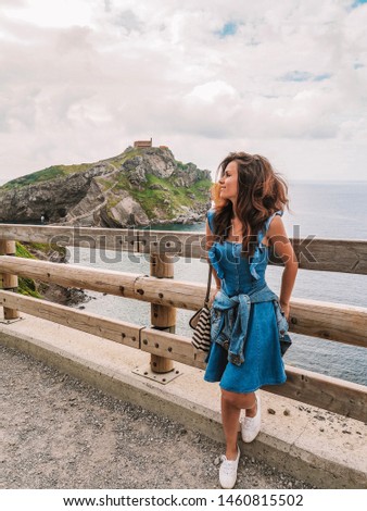 Young girl in blue dress with long hair and earrings rises on the island Gaztelugatxe on the coast of the Bay of Biscay, Spain, Basque Country