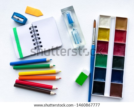 Flat lay of stationery for school and creativity, drawing and crafts (watercolor paints, colored pencils, notebook, hourglass) is on the right against a white textured rough watercolor paper 