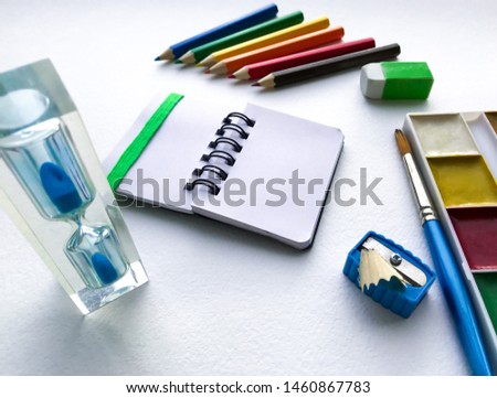 Close-up of hourglass and stationery for school and creativity, drawing and crafts (watercolor paints, colored pencils, eraser, sharpener) against a white textured rough watercolor paper