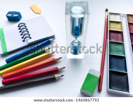 Close-up of hourglass and stationery for school and creativity, drawing and crafts (watercolor paints, colored pencils, eraser, sharpener) against a white textured rough watercolor paper