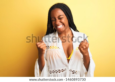 African american woman on vacation wearing bikini and shirt over isolated yellow background very happy and excited doing winner gesture with arms raised, smiling and screaming for success.