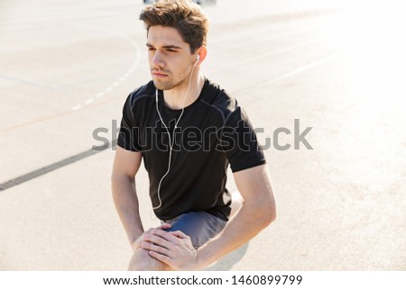 Image of attractive man in sportswear wearing earphones training on sports ground during morning workout outdoors