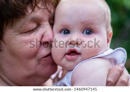 Little cute baby girl on her grandmother's hands. Grandma is holding and kissing a baby. Summer garden, green trees, heat.