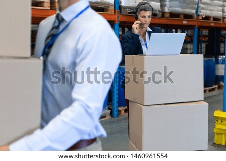 Front view of mature Caucasian female manager talking on headset while working on laptop in warehouse. On the foreground Caucasian male holding cardboard boxes. This is a freight transportation and