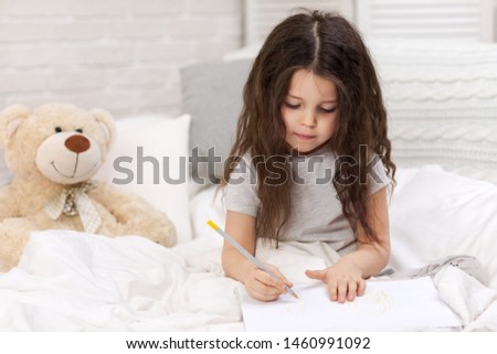 cute little girl with teddy bear drawing pictures while lying on bed. Kid painting at home