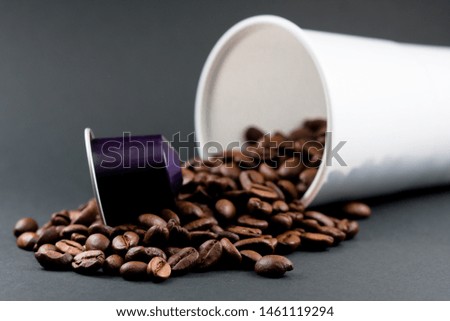 Plastic cup of white coffee lying, across a black background, with brown coffee beans inside the glass, and a capsule on top of the coffee beans. Cafeteria and food.