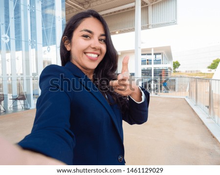 Joyful friendly business professional choosing you. Happy beautiful young Latin woman in office suit taking selfie and pointing index finger at camera. Selection concept