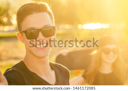 cool guy and a lovely girl in sunglasses, jeans and a black T-shirt skateboarding holding hands photographed take pictures selfie on phone in the summer park