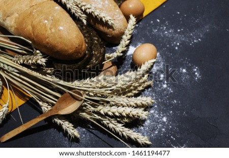 White bread baguette, black bread with cereals, wheat ears, chicken eggs, wooden spoon, brown cutting board and yellow cloth on a gray textured surface covered with flour