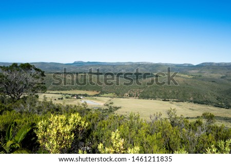 A view from the top of a hill. Bushes in the foreground with mountains and farmland in the background with bright blue sky on a cloudless sunny day.