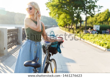 Photo of curly blonde looking at side in denim skirt standing next to bike on bridge in city