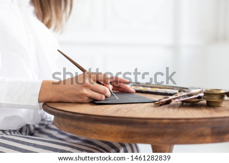Woman calligrapher is holding a pen with ink and signs a card in handwritten font. Close-up. Soft focus.