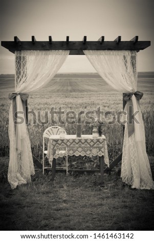 
Rustic arbor decoration with white curtains and burlap ties and a wooden table chairs and lace tablecloth at a vintage country wedding with the wedding aisle in front of a wheat field on a farm