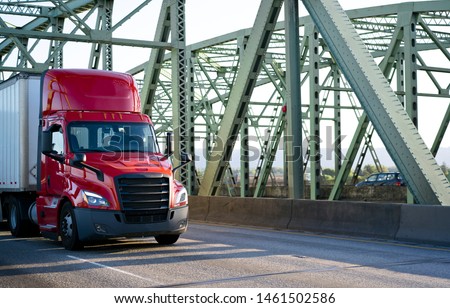 Big rig bright red day cab powerful bonnet semi truck transporting commercial cargo in dry van semi trailer with aerodynamic skirt driving on the metal truss Columbia River Interstate drawbridge Royalty-Free Stock Photo #1461502586