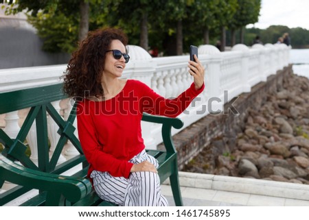 Stylish beautiful curly woman in sunglasses doing selfie on a smartphone, seated on a green bench in the park.