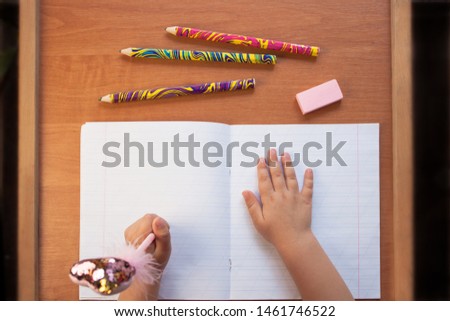 A child holds a pen in the left hand and writes in an empty notebook on a wooden desk. Stationery, colour pencils and eraser. Back to school concept. Top view, copy space.