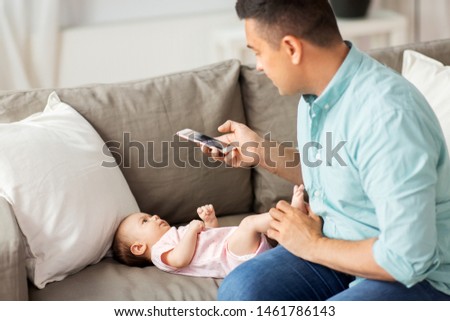 family, technology and fatherhood concept - middle aged father with smartphone taking picture of his little baby daughter lying on sofa at home