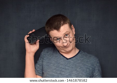 Studio close-up portrait of funny caucasian fair-haired man, wearing t-shirt, scratching head using phone like being in amusing and silly confusion after ridiculous conversation, over gray background