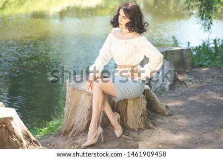 cute girl in a white summer dress tired of the sun and sat on a large stump near the lake in the shade of trees