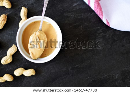 A bowl full of peanut butter lies on a dark marble surface, with a spoon on it and some peanuts beside it - concept with delicious sweet and savory peanut butter