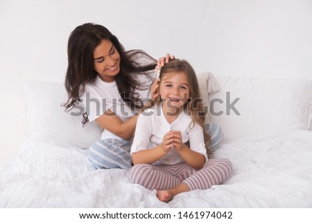 Happy family - smiling mother braids her daughter's hair and coif her in bed early in the morning isolated
