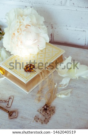 A beautiful white peony lies on a book with Golden pages. Near are delicate petals vintage keys and embellishment on the neck. Vintage background in light white and beige.