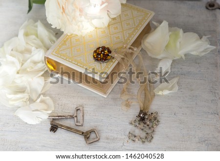 A beautiful white peony lies on a book with Golden pages. Near are delicate petals vintage keys and embellishment on the neck. Vintage background in light white and beige.