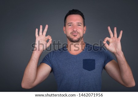 Glad attractive man shows ok sign with both hands as expresses approval, has cheerful expression. Photo of beautiful female has appealing appearance, being optimistic. Standing against gray wall.