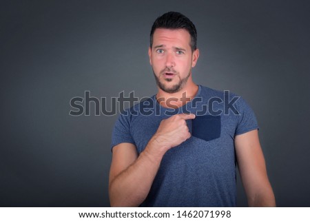 Surprised handsome man student being in stupor shocked, has astonished expression pointing at herself with finger saying: Who me? Isolated over gray background.