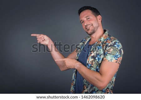 Positive attractive young male wearing Hawaiian flowers shirt, points aside with cheerful  expression with mouth opened, shows something amazing standing indoors. Advertisement concept.