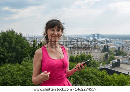Beautiful woman with dark hair wearing pink dress holding smartphone in her hands having earphones in her ears listening to the music, looking at the camera and smiling. Outdoors.