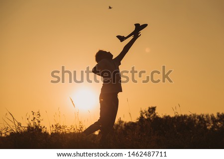 Black silhouette of cute happy cheerful child playing at grassy hill at countryside holding big toy plane in hand. Boy has fun during sunset time in evening. Horizontal color photography.