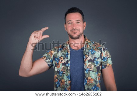 Blond European man wearing Hawaiian flower shirt over isolated background gesturing with hand showing small size, measure symbol. Smiling looking at the camera. Measuring concept.