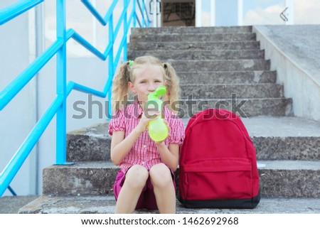 Back to school. Beautiful blond schoolgirl drinking water from a bottle with red backpack and books outside the primary school , education concept
