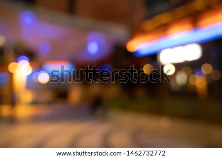 The picture is blurred in a shopping mall at night that turns on the lights beautifully. There are people who are happy shopping. The color are red-blue and lively