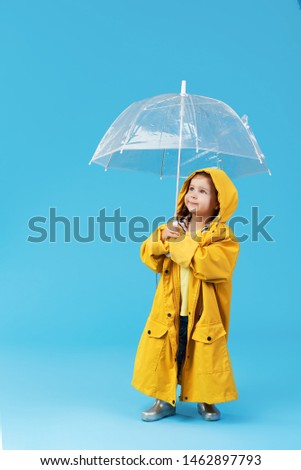 Happy funny child with transparent umbrella posing on blue studio background. Girl is wearing yellow raincoat and rubber boots. Holds a vintage travel suitcase