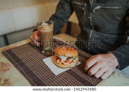 Food photography. Hot cappuccino with hamburger at the table in fast food cafe. Cheeseburger and coffee on the table, drink is containing caffeine.