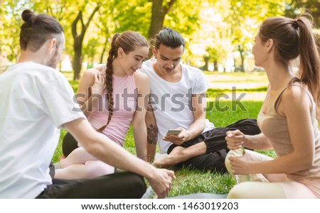 Portrait Of Cheerful Young Friends Looking At Smart Phone Sitting On A Grass In Park